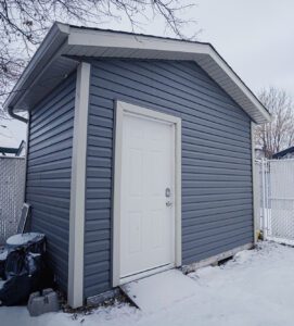 A small grey shed with a white door on a snowy ground.